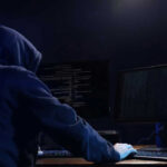 Hooded figure using dual monitors in dark room.