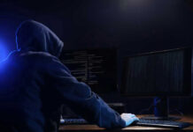 Spyware Scandal Explodes – Journalists Targeted Hooded figure using dual monitors in dark room.