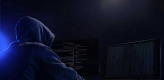 Hooded figure using dual monitors in dark room.