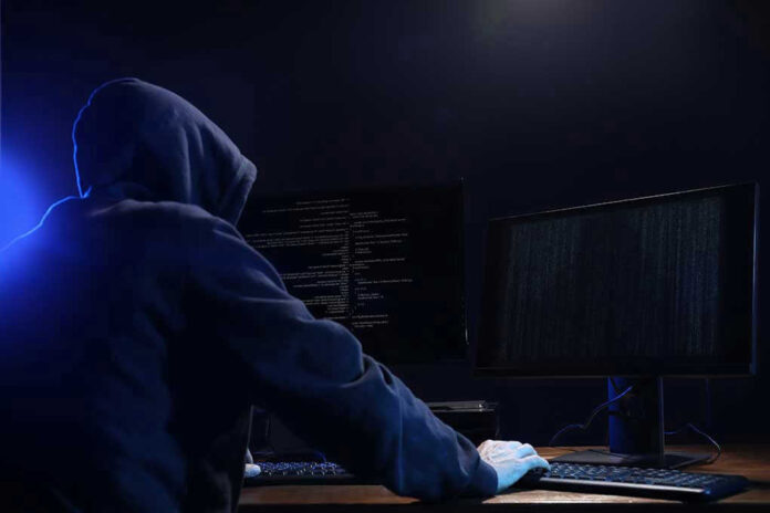 1708895812 Hooded figure using dual monitors in dark room.