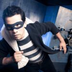 A masked thief in a striped shirt holding a bag while emerging from a bank vault