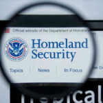 Migrant Stipend Sparks Outrage: Holiday Bribe? Magnifying glass showing Homeland Security website.