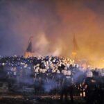 A large crowd of police officers in helmets facing a smoky protest scene at night