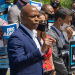 Mayor’s First Day Bombshell: Adams’ Directives Erased Man speaking at campaign event with supporters behind him.