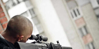 Person aiming a rifle from a building balcony.