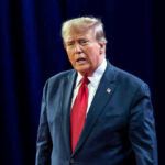 Trump Drops Endorsement THREAT Over Vote Man in suit with red tie speaking on stage.