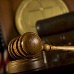 Fauci Advisor INDICTED — COVID Cover-Up Exposed A wooden gavel resting on a desk in a courtroom with an American flag in the background