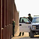 Surprise Exiles: Asylum Seekers’ MYSTERIOUS Mass Exit Border patrol agents near a tall metal fence.