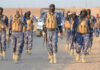 Camouflaged soldiers wearing masks and vests walk in desert.