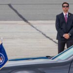 Kash Patel’s Quiet Funeral Turns into Public Spectacle A security officer in a suit stands guard near a presidential vehicle at an airport