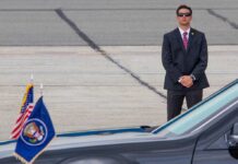 Kash Patel’s Quiet Funeral Turns into Public Spectacle A security officer in a suit stands guard near a presidential vehicle at an airport