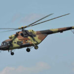 Apache Down Near Hormuz — What Hit It? Camouflage military helicopter in flight against blue sky.