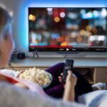 Person sitting on a couch watching a movie on a TV with popcorn in hand