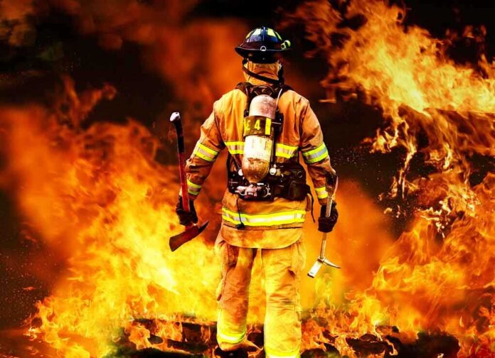 shutterstock_80081197 Firefighter standing in front of a large fire, equipped with tools