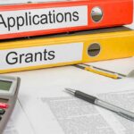 UC Riverside Picks Winners — US Citizens Last? Two binders labeled Applications and Grants on a desk with a calculator and papers
