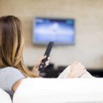 A woman sitting on a sofa, holding a remote control and watching television