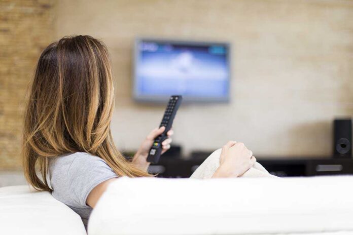 Young,Woman,Watching,Tv,In,The,Room A woman sitting on a sofa, holding a remote control and watching television