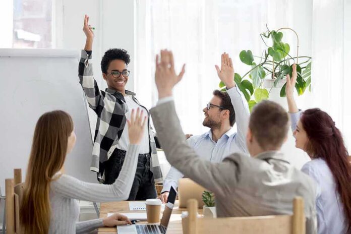 shutterstock_1296003193 Team in meeting raising hands around a smiling presenter