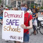 Protest Uproar Hijacks Mourning – Teacher Named Person wearing mask holds protest sign reading Demand SAFE Schools